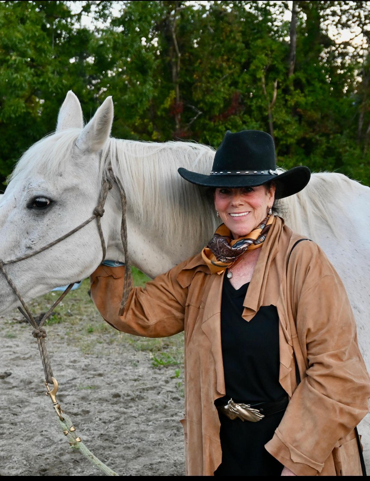 Barbara Newman wearing a black cowgirl hat, standing beside a white horse